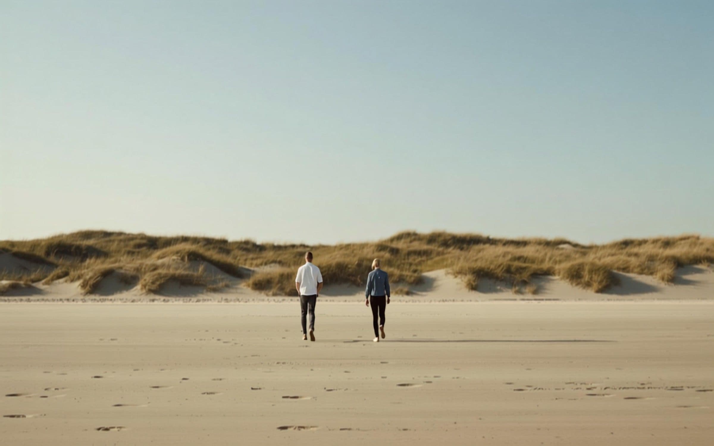 Twee mensen in gesprek, wandelend over een breed Nederlands strand met duinen in warm ochtendlicht